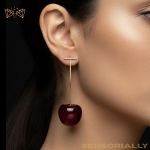 May include: Gold-coloured cherry earrings. The earrings have a small horizontal bar that attaches to a long, thin vertical bar. A dark red cherry hangs from the bottom of the vertical bar. The earrings are worn on the ear of a person.