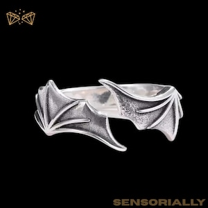 Może przedstawiać: Srebrny pierścionek z motywem skrzydła nietoperza. Pierścionek posiada dwa szczegółowe skrzydła, które owijają się wokół palca. Pierścionek jest umieszczony na czarnym tle. Słowo "SENSORIALLY" znajduje się na dole obrazu.