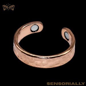 May include: A copper-colored ring with a hammered texture and two embedded magnets. The ring has an open design and is set against a black background. The word "SENSORIALLY" is at the bottom of the image.
