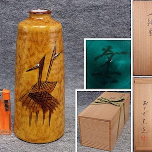 May include: A cylindrical ceramic vase with a mottled yellow-brown glaze featuring a dark brown painted crane design. The vase is shown next to an orange lighter, a close-up of a dark mark, and a wooden storage box with Japanese calligraphy.