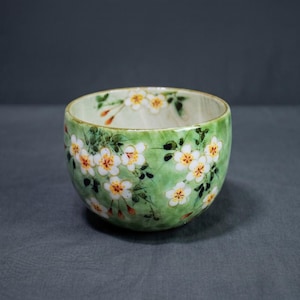 Puede incluir: Un cuenco de cerámica pintado a mano con un fondo verde y un diseño floral blanco. El cuenco tiene forma redondeada y un acabado liso y brillante. Las flores tienen centros amarillos y detalles en naranja.