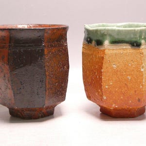 May include: Two hexagonal ceramic cups with textured surfaces. The left cup features a dark brown and black glaze with a rough finish. The right cup displays a light orange and tan body with a distinct green glaze dripping along the top rim.