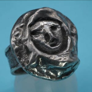 May include: A silver-toned ring featuring a sculpted face design. The face has detailed features and is surrounded by a textured, organic shape. The ring band is simple and smooth, contrasting with the detailed face. The ring is set against a light blue background.