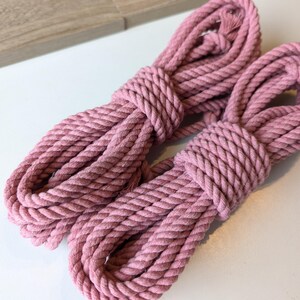 Puede incluir: Dos bobinas de cuerda de algodón trenzado en color rosa empolvado. Cada tramo de cuerda está cuidadosamente enrollado en forma ovalada con textura de fibra visible y extremos deshilachados. La cuerda presenta una construcción trenzada.