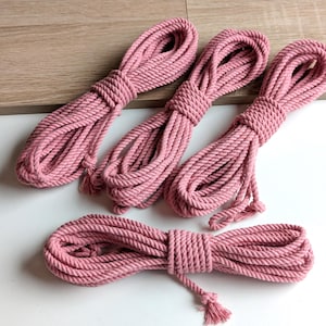 Puede incluir: Cuatro bobinas de cuerda de algodón de color rosa empolvado, cada una enrollada cuidadosamente y asegurada con una vuelta central. Las cuerdas presentan una textura trenzada y están dispuestas con tres bobinas verticalmente y una horizontal.