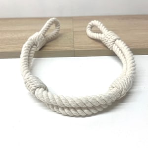 Può includere: Maniglia in corda color crema con estremità ad anello, adatta a varie applicazioni. La corda è strettamente intrecciata, creando un aspetto spesso e resistente. La maniglia è posizionata su una superficie di legno chiaro.