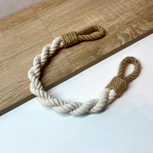 May include: A twisted rope handle with loops at each end. The handle is made of off-white and tan rope. The rope is lying on a light wood surface and a white surface.
