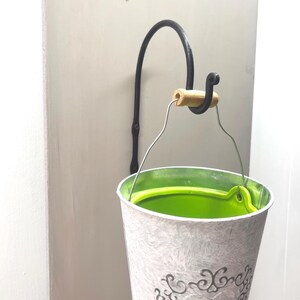 Pode incluir: Um suporte de planta de parede de metal preto com um botão de madeira e um balde de metal branco com um forro verde.