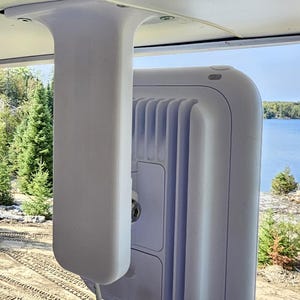 Pode incluir: Um dispositivo retangular branco com uma borda curva e uma série de cristas verticais na parte traseira. É montado em uma estrutura de suporte branca, com um cabo que se estende da parte inferior. O fundo mostra um lago e árvores.