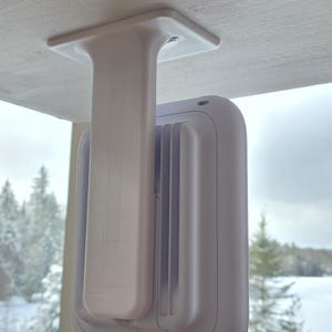 May include: A white, rectangular ceiling mount with a vertical support and a rounded, rectangular device attached. The device has a series of vertical ridges on its side. The mount is attached to a white wooden surface.