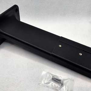 May include: Black plastic wall mount with two screw holes and a slot for a cable. The mount is designed to hold a device or object.