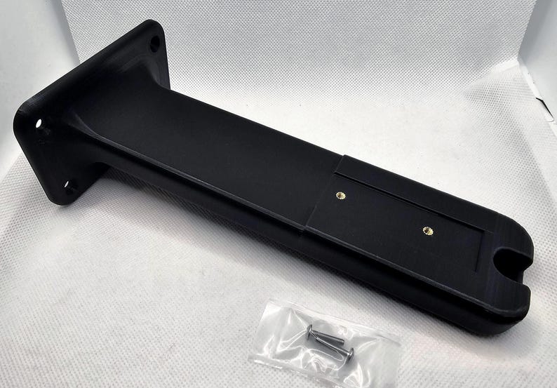 May include: Black plastic wall mount with two screw holes and a slot for a cable. The mount is designed to hold a device or object.