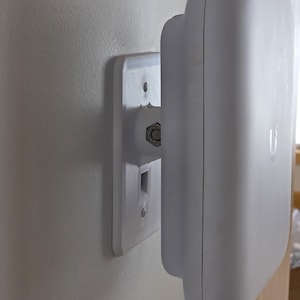 Pode incluir: Um dispositivo de rede branco, de forma retangular arredondada, montado numa placa branca. A placa está fixada a uma parede de cor clara. O dispositivo tem um conector central e dois orifícios para parafusos para montagem.
