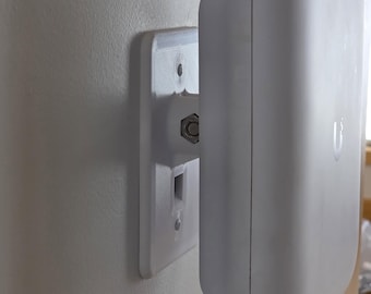 May include: A white, wall-mounted network device with a rounded rectangular shape. It is attached to a white mounting plate with a central connector and two screw holes. The device is mounted on a light-colored wall.