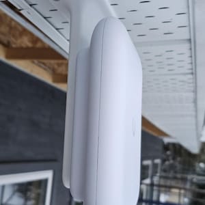May include: A white wireless router mounted on a white wall with a white bracket. The router has a rounded rectangular shape and a small logo on the front.