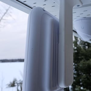 May include: A white security camera mounted on a white bracket attached to a ceiling. The camera is facing downwards and is positioned in front of a window with a snowy landscape in the background.
