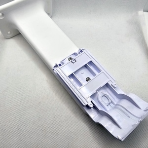 May include: White plastic wall mount with a slot for a device. The mount has a white plastic base and a white plastic arm that extends out from the base. The arm has a slot for a device to be inserted into it. The mount is designed to be attached to a wall.