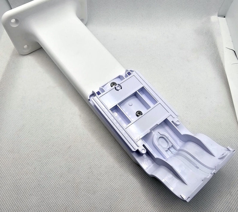 May include: White plastic wall mount with a slot for a device. The mount has a white plastic base and a white plastic arm that extends out from the base. The arm has a slot for a device to be inserted into it. The mount is designed to be attached to a wall.