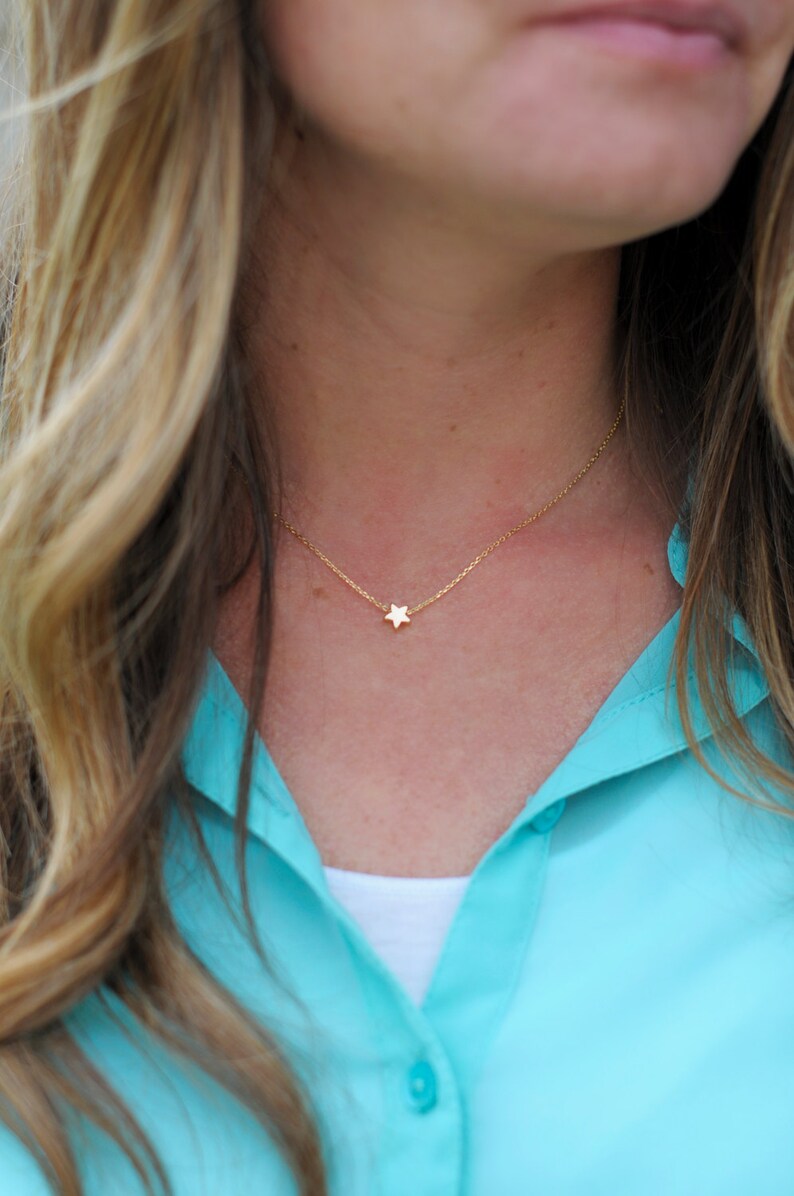 May include: A gold chain necklace with a small star pendant. The necklace is worn by a person with long blonde hair and a turquoise shirt.