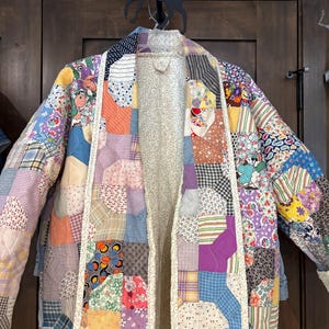 May include: A patchwork jacket with a variety of fabric patterns and colors, including floral, geometric, and solid patches. The jacket is open, revealing a patterned lining. It is hanging on a wooden door.