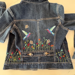 May include: A dark wash denim jacket with colorful embroidered hummingbirds, butterflies, and flowers. The jacket features a classic collar, button closures, and pockets. The embroidery includes shades of blue, green, red, and yellow.