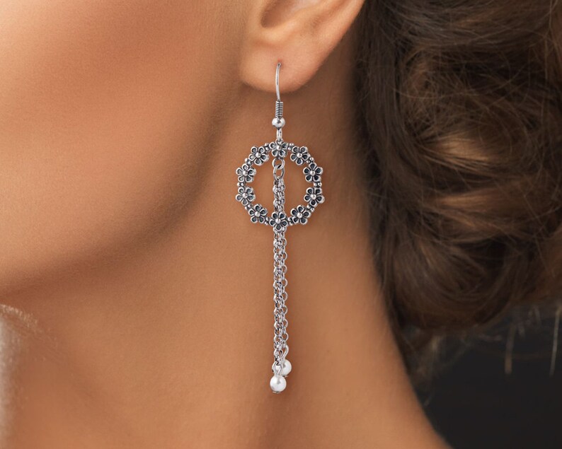 May include: Silver dangle earrings with a circle design featuring small flower accents and a chain with two pearl-like beads at the bottom.