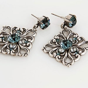 Peut inclure: Une paire de boucles d'oreilles pendantes en argent avec un design carr&eacute;. Les boucles d'oreilles pr&eacute;sentent un cristal bleu au centre de chaque carr&eacute;, entour&eacute; de petits cristaux.
