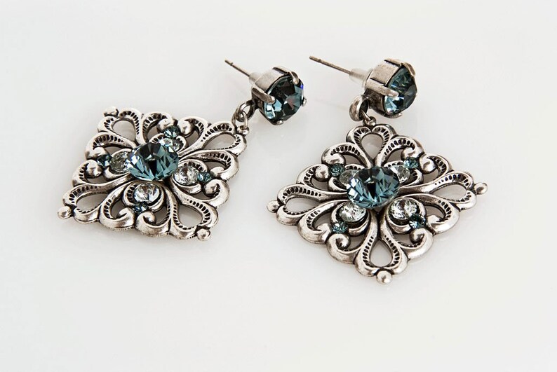 Peut inclure: Une paire de boucles d'oreilles pendantes en argent avec un design carr&eacute;. Les boucles d'oreilles pr&eacute;sentent un cristal bleu au centre de chaque carr&eacute;, entour&eacute; de petits cristaux.