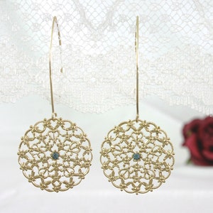 May include: A pair of gold filigree earrings with a blue gemstone in the center of each circle. The earrings are hanging from a white lace background.