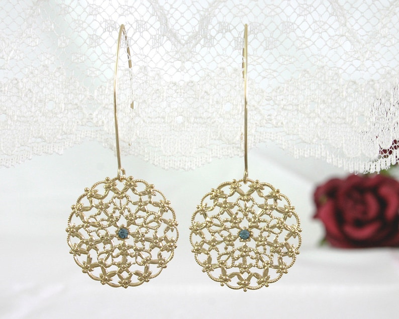 May include: A pair of gold filigree earrings with a blue gemstone in the center of each circle. The earrings are hanging from a white lace background.