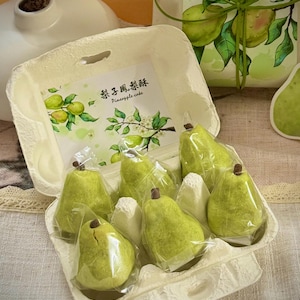 May include: A carton of pear-shaped pastries, individually wrapped in clear plastic. The pastries are light green with a brown stem. The carton has a printed label with text and a watercolor illustration of pears. A gift box with a pear illustration is in the background.