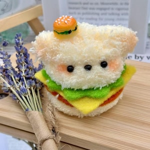 May include: A plush toy shaped like a sandwich with a cute bear face. The sandwich has layers of yellow, green, and red, topped with a miniature burger. The bear has black eyes and pink cheeks. A bundle of lavender is in the background.