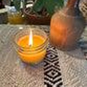 Puede incluir: Una vela naranja encendida en un tarro de vidrio transparente con un patrón decorativo en el fondo. La vela está sentada sobre un mantel estampado.