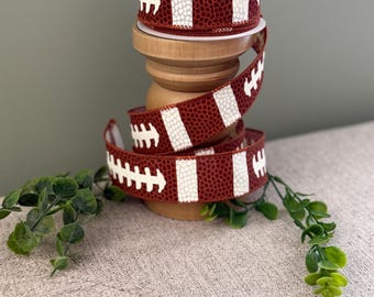 May include: A spool of burgundy ribbon with a football pattern, featuring white stripes and stitching details. The ribbon is displayed on a wooden stand, with a sprig of green leaves. Perfect for crafting and sports-themed projects.