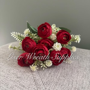May include: A cluster of vibrant red artificial flowers, complemented by green foliage and tiny white blooms. The arrangement rests on a grey surface. The text "Liser Wreath Supplies" is visible.