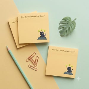 May include: Square light yellow sticky notes featuring the text How Can I Get More Stuff Done? and a graphic of a black cat wearing a headband with lightbulb icons. Included are copper-colored metal paper clips and a light green pencil.