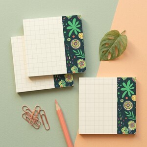 May include: Square sticky note pads featuring a white grid pattern on the left and a dark blue vertical strip on the right decorated with colorful botanical and circular motifs, accompanied by copper-colored paper clips and a peach-colored pencil.