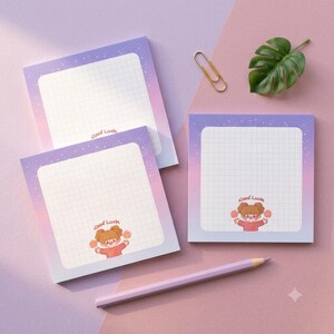 May include: Three square sticky notes with a purple and pink gradient background, featuring a white grid center and the text Good Luck. One note shows a cartoon character with brown hair holding flowers. Includes a gold paper clip and a light purple pencil.