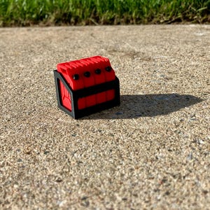 May include: A small, red, rectangular object with black accents and four black dots on top. The object is sitting on a concrete surface, casting a shadow. The object appears to be a decorative item or a small container.