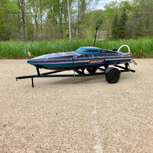 May include: A black and teal remote-controlled boat with the word "SHREDDY" on the side, mounted on a black trailer. The boat has a lightning bolt design and a white propeller. The trailer has black wheels and a hitch.