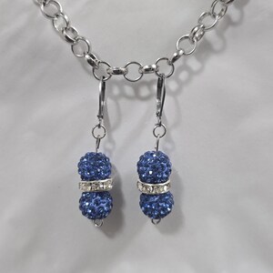 Op de afbeelding: Hangende oorbellen met een zilveren ketting. Elke oorbel heeft een cluster van kleine, donkerblauwe, sprankelende kralen, gescheiden door een zilveren band met heldere strass-steentjes. De oorbellen hangen aan zilveren oorhaaksluitingen.