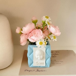 Könnte beinhalten: Hellblaue quadratische Vase mit Steppmuster, gefüllt mit rosa und weißen Blumen. Ein weißes Relief eines Babygesichts und der Name "Sam" befinden sich auf der Vorderseite. Die Vase steht auf einem Regal mit einem Buch.