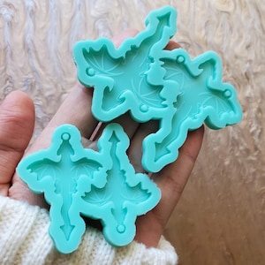 May include: Two turquoise silicone molds for making resin dragon charms. The molds are in the shape of dragons with wings and tails. The molds are held in a person's hand.