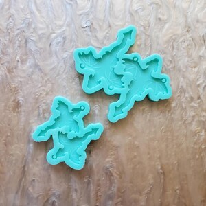 May include: Two turquoise silicone molds for making resin dragon charms. The molds are shaped like dragons with wings and tails. The larger mold has two dragons, while the smaller mold has one dragon.