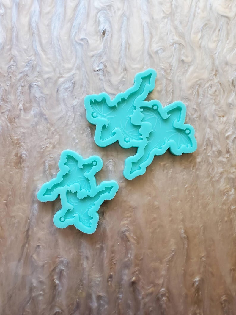 May include: Two turquoise silicone molds for making resin dragon charms. The molds are shaped like dragons with wings and tails. The larger mold has two dragons, while the smaller mold has one dragon.