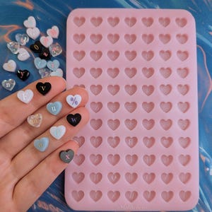 May include: A rectangular pink silicone mold featuring a grid of heart-shaped cavities, each embossed with a letter or symbol. Beside the mold are several small, individual heart-shaped pieces in various colors, including white, black, and blue, each marked with a letter or symbol.