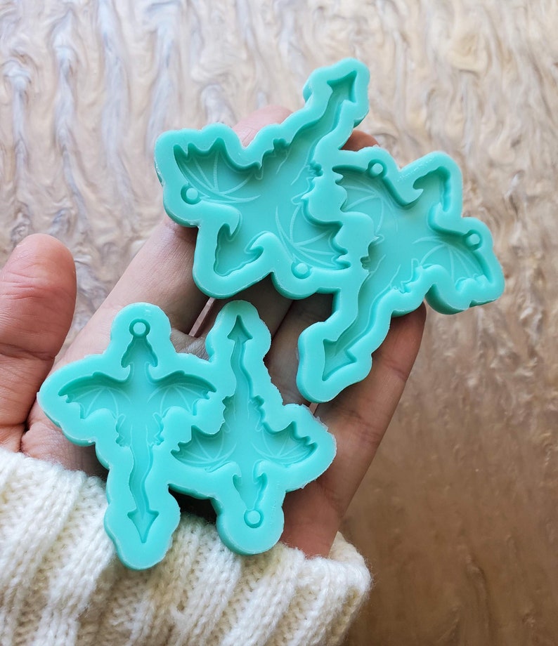 May include: Two turquoise silicone molds for making resin dragon charms. The molds are in the shape of dragons with wings and tails. The molds are held in a person's hand.