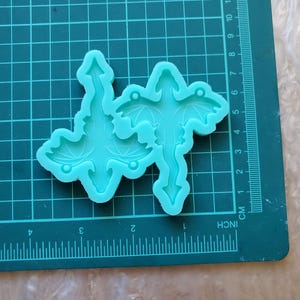 May include: A light blue silicone mold for making dragon-shaped resin ornaments. The mold has two dragon shapes, each with wings and a tail.