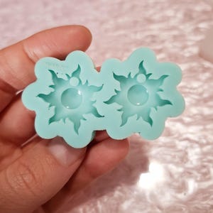 Puede incluir: Un molde de silicona azul claro con dos cavidades en forma de sol. Cada cavidad tiene una hendidura redonda en el centro.