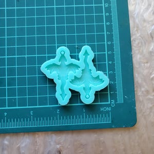 May include: Two teal blue silicone molds of a dragon with wings outstretched and a tail that ends in a point. The molds are on a green cutting mat with inch and centimeter markings.